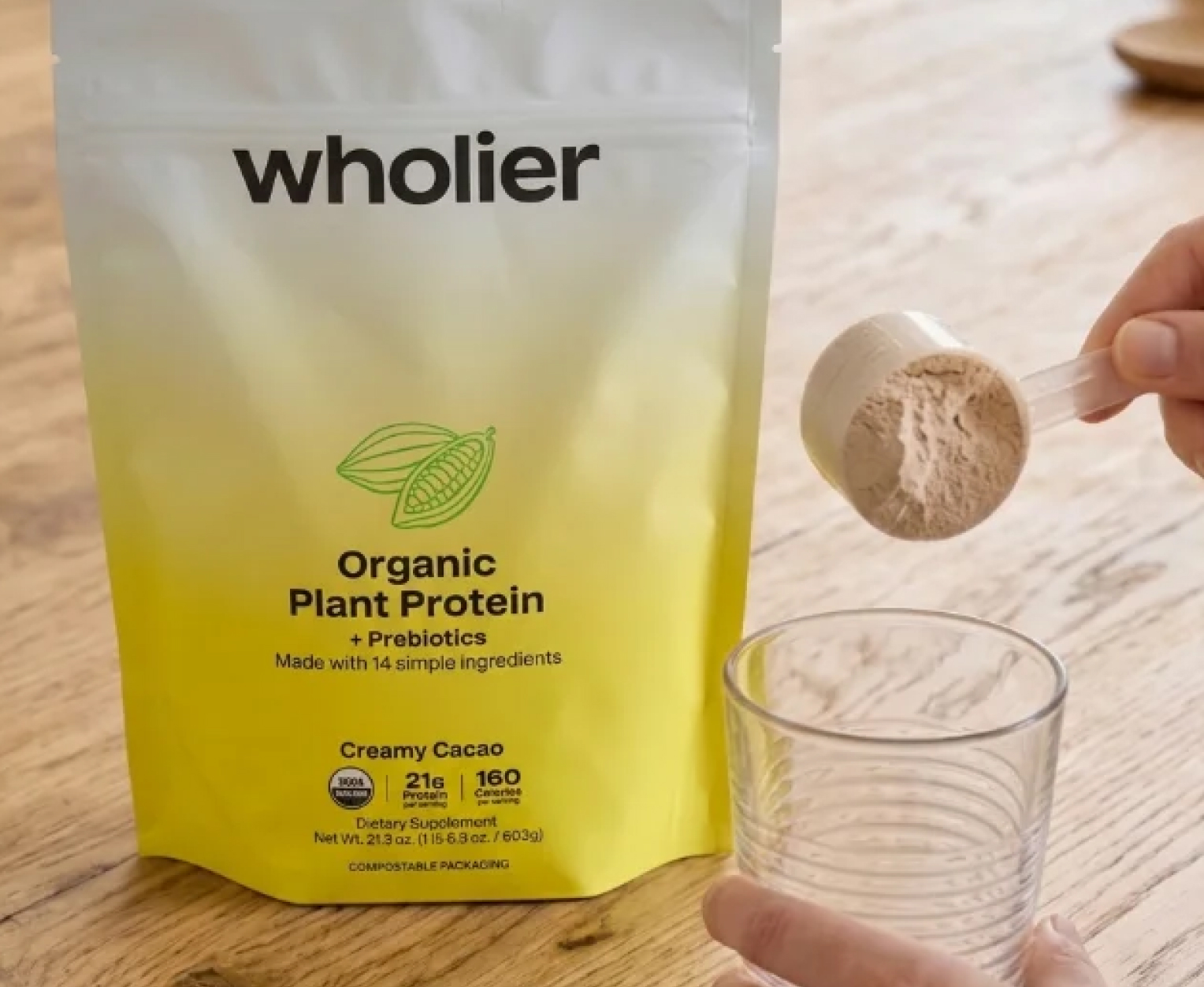 A person holds a scoop of wholier Organic Plant Protein powder over a glass, next to the product pouch.