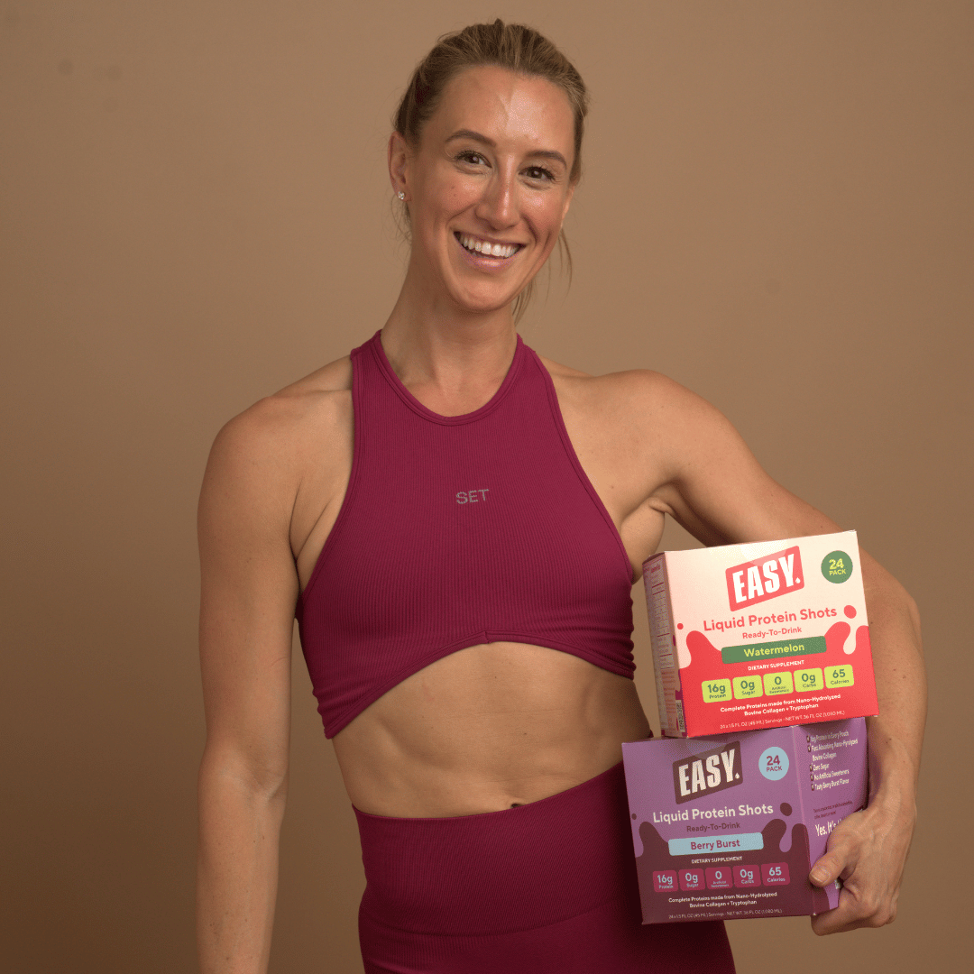 Person in workout attire holding protein shot boxes, smiling.