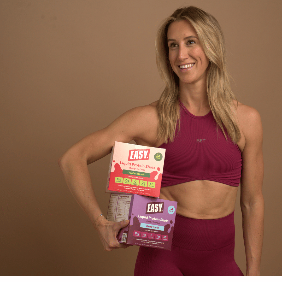 Woman in workout attire holding protein shot boxes, smiling.