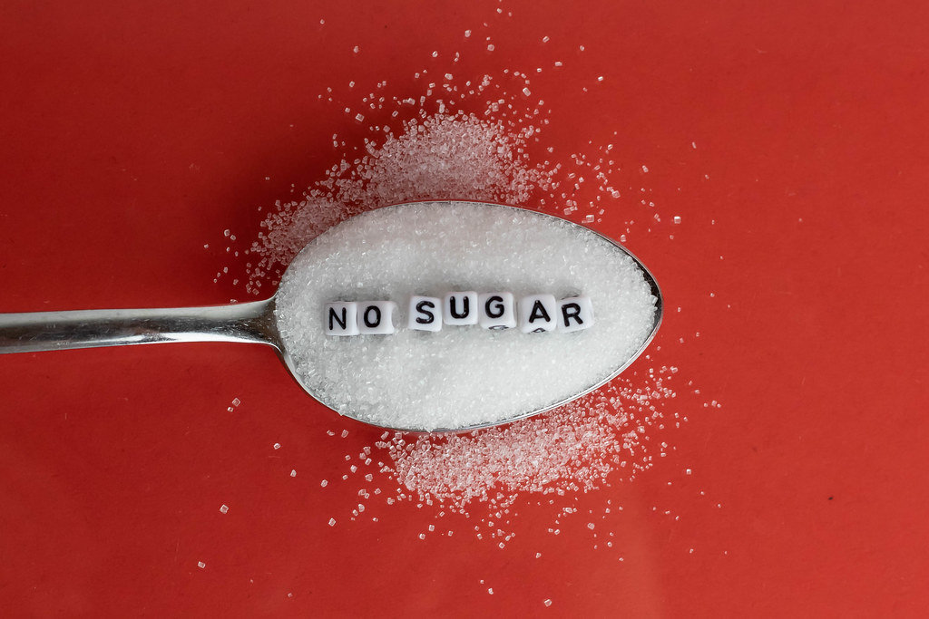 Spoon with sugar and 'NO SUGAR' letter beads on red background.