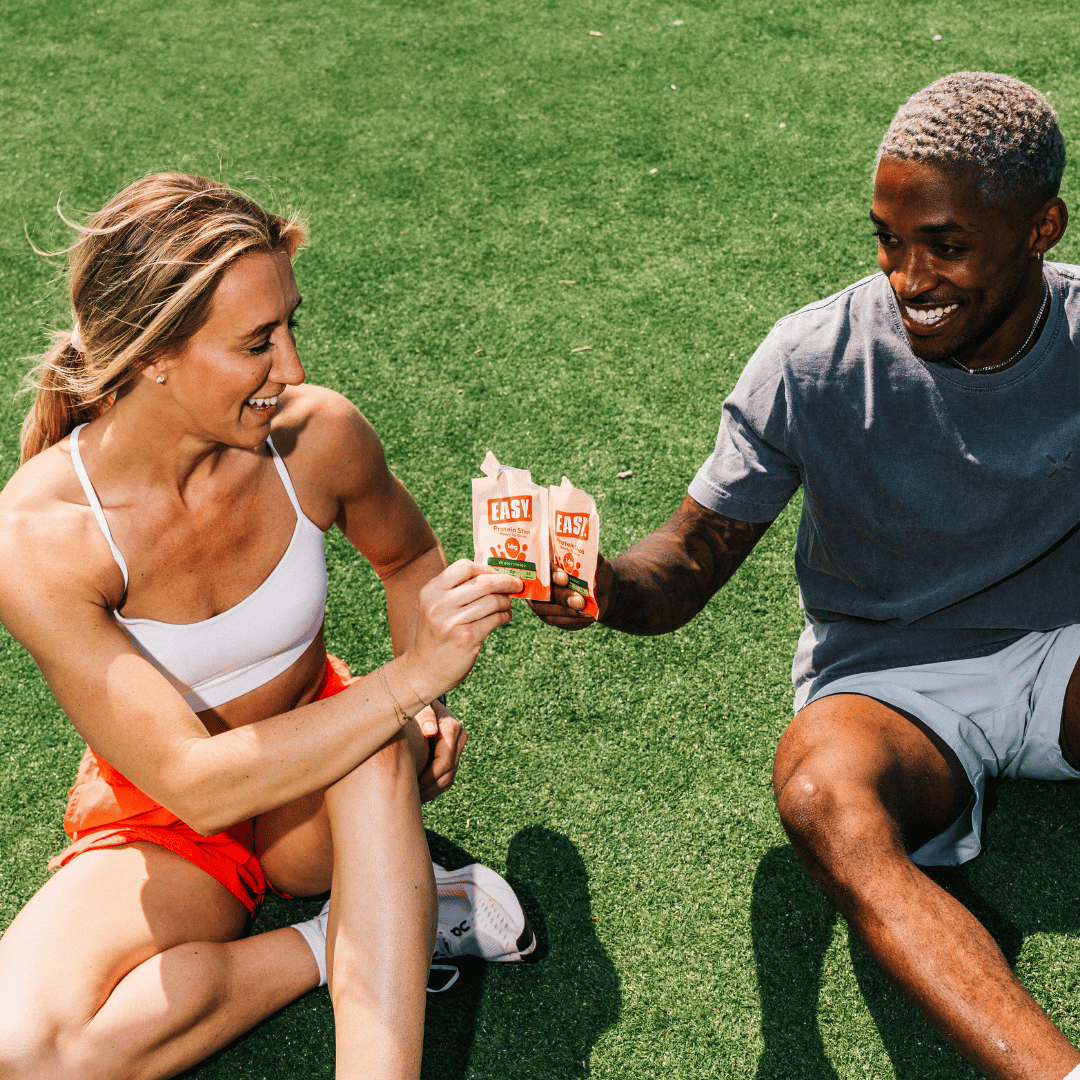 Two people sitting on grass, holding snack packs and smiling.