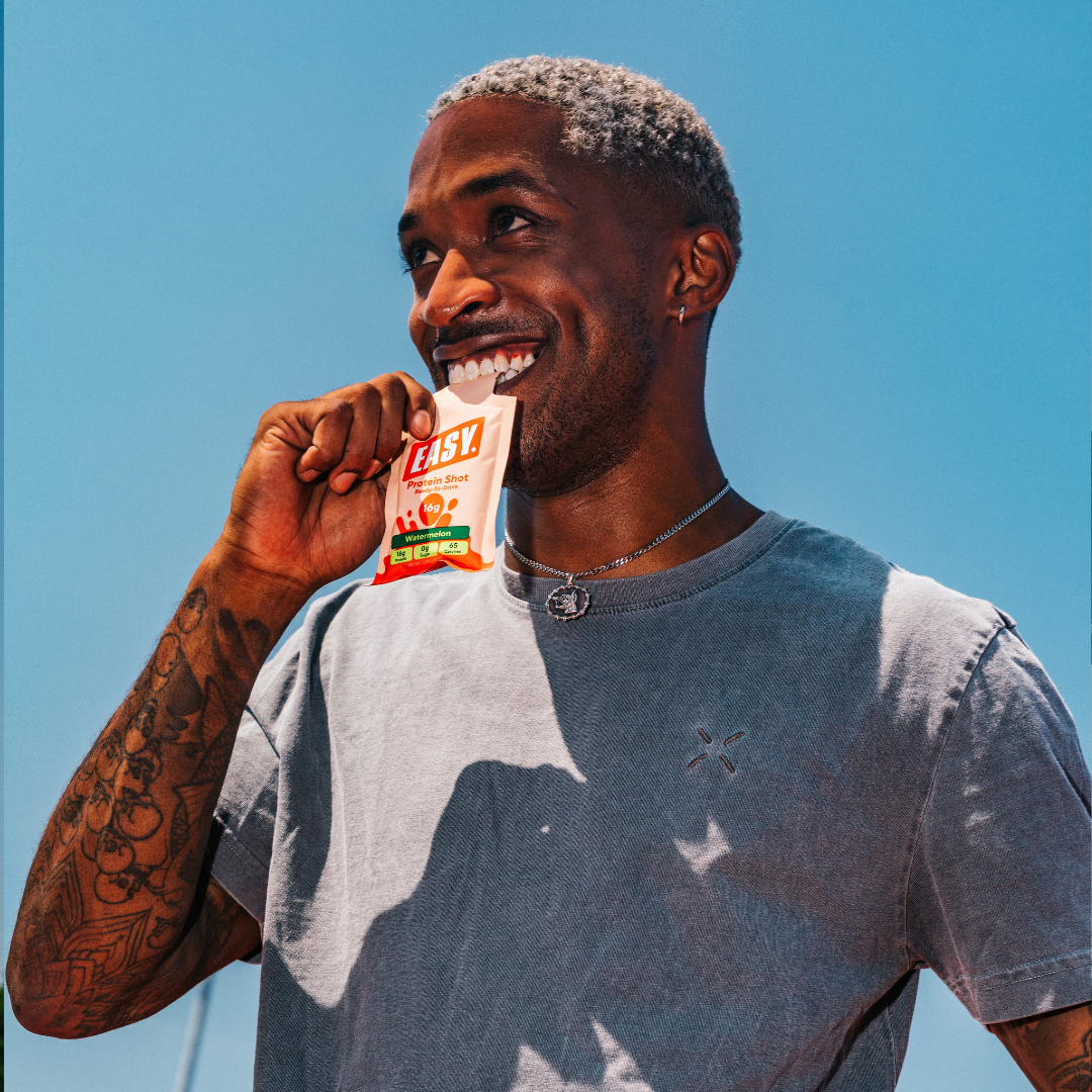 Person smiling, holding a watermelon-flavored protein shot against a clear blue sky.