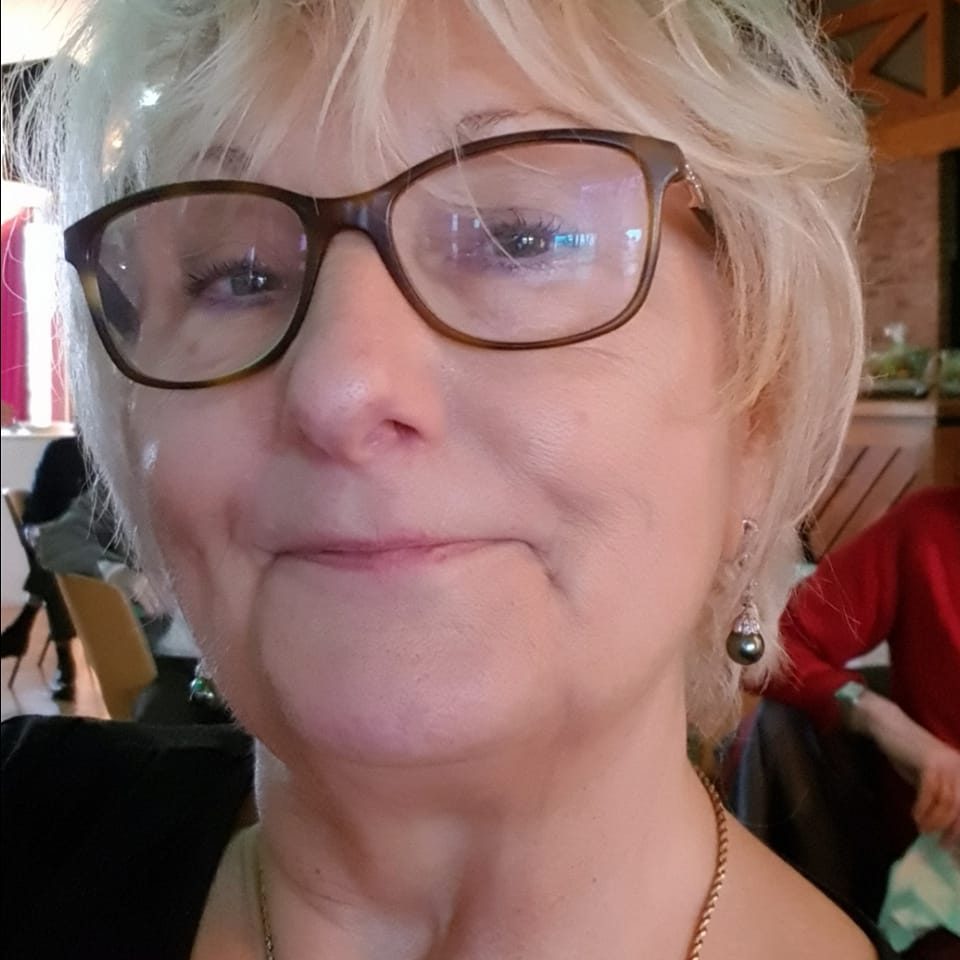 An elderly person wearing glasses and earrings, smiling slightly indoors.