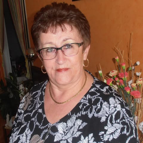 An older woman with glasses stands beside flowers, wearing a floral patterned blouse.