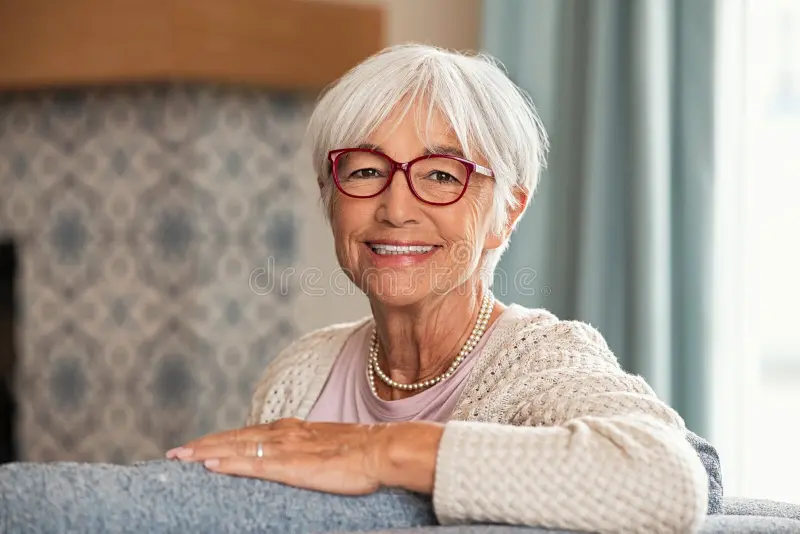Smiling older woman with red glasses sitting on a couch at home.