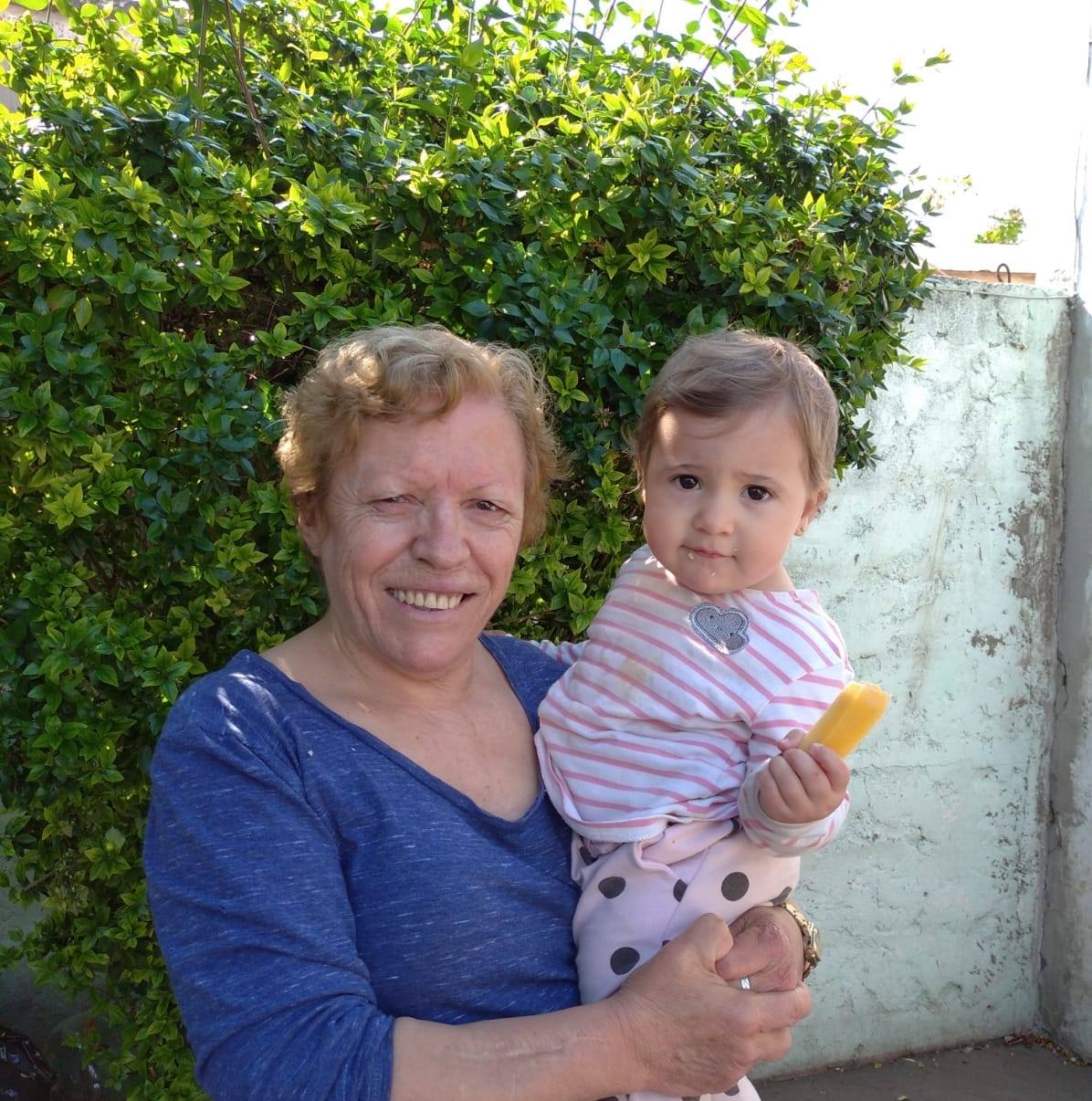 Elderly woman holds a young child with a popsicle, standing in front of green foliage.
