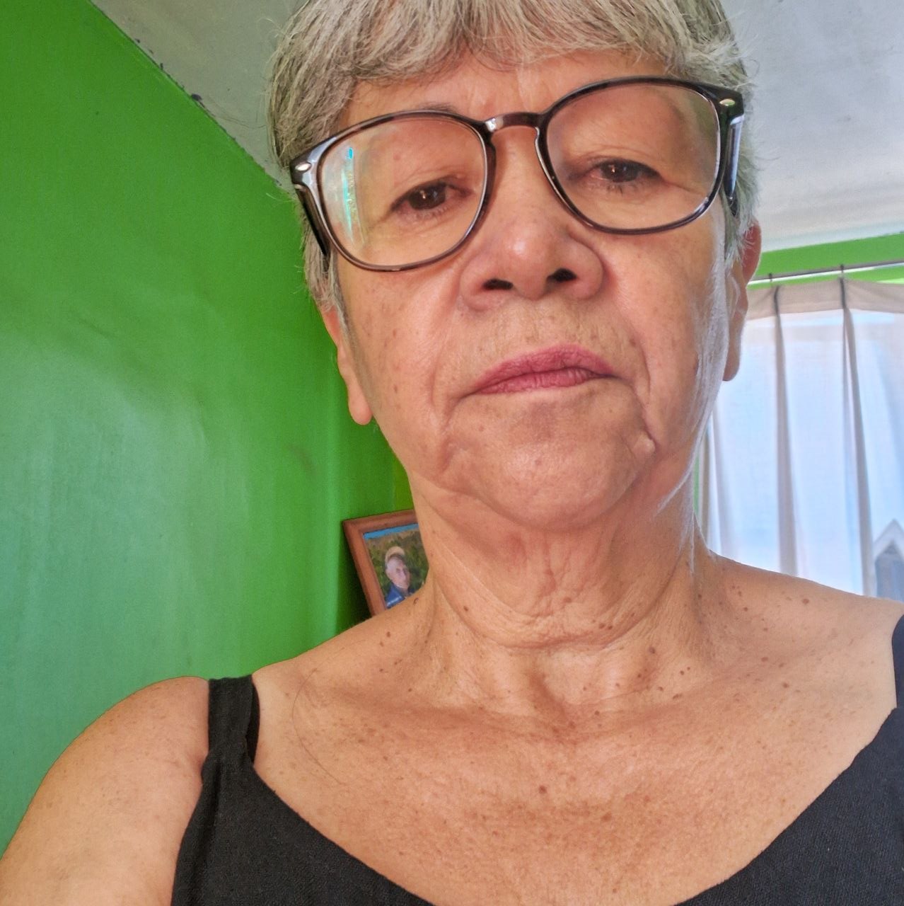 Elderly person with glasses indoors, green walls, framed photo in background.