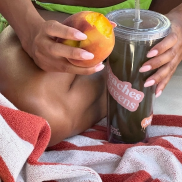 Person holding a peach and a cup labeled 'Peaches Greens' with a striped towel nearby.