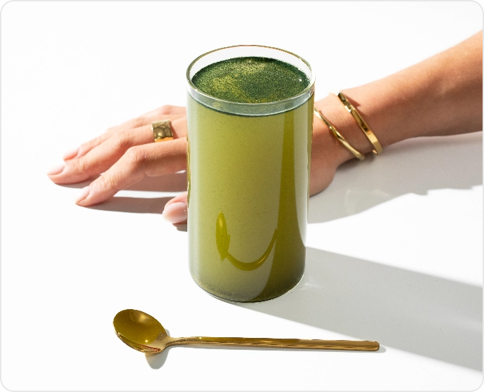 A glass of green liquid with a gold spoon and a hand wearing jewelry.