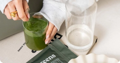 Person stirring a green drink next to a glass of water and a greens packet.
