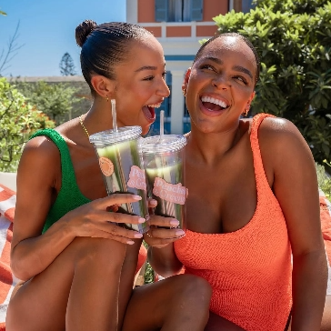 Two women in swimsuits laughing and holding drinks outdoors.
