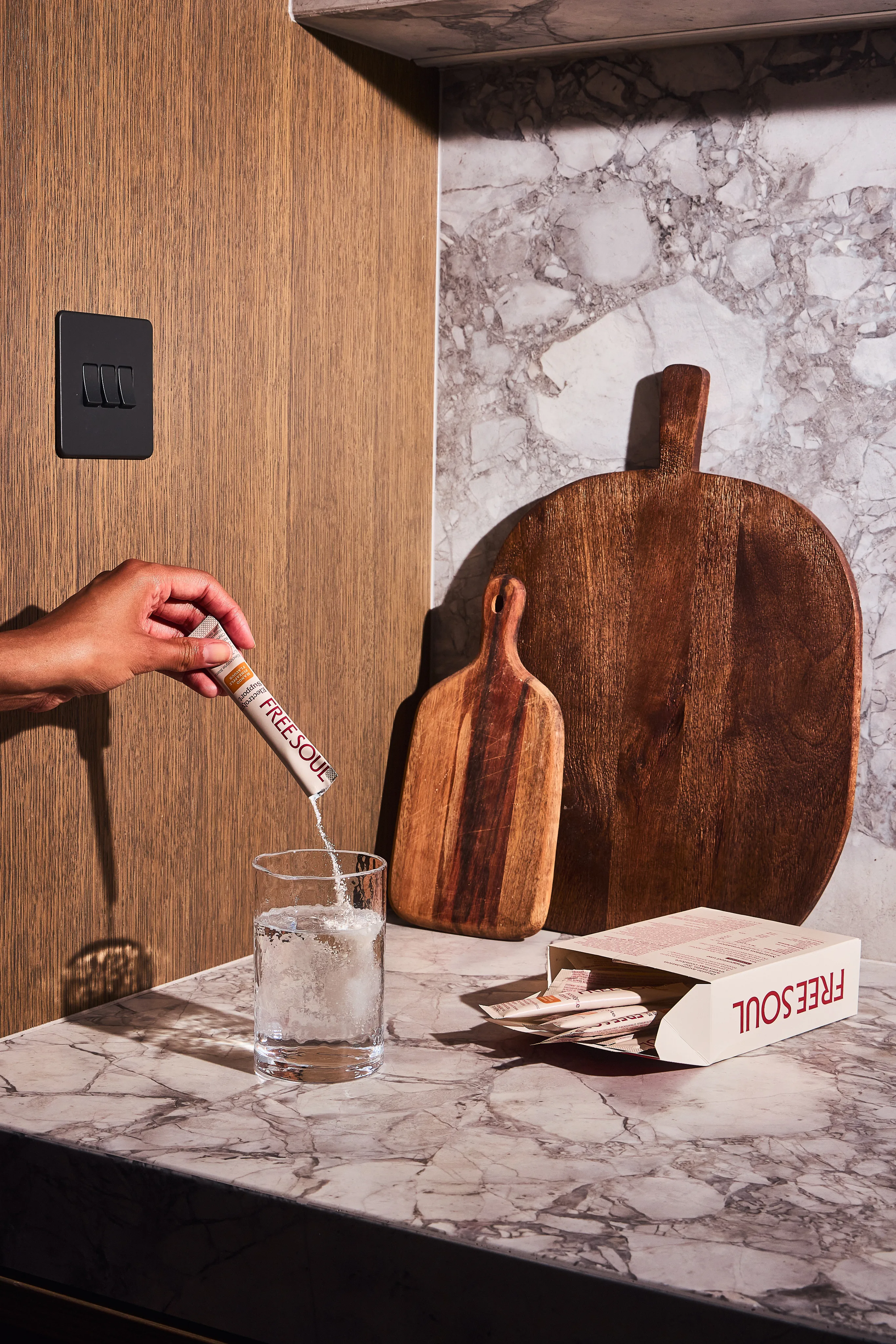 A hand pours a Freesoul supplement sachet into a glass of water on a marble kitchen counter.