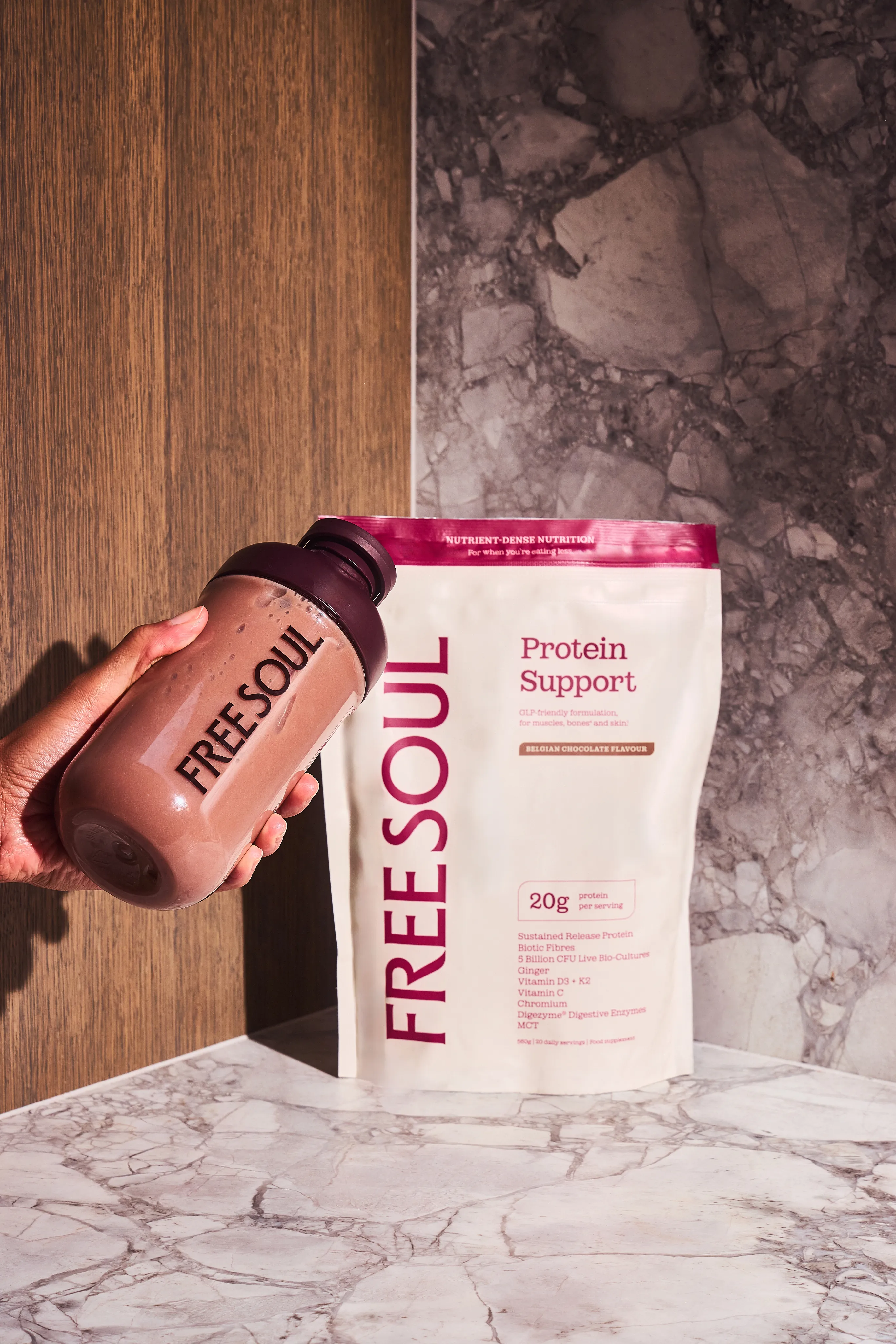 A hand holds a Free Soul protein shake next to a pouch of Belgian Chocolate flavour protein powder.