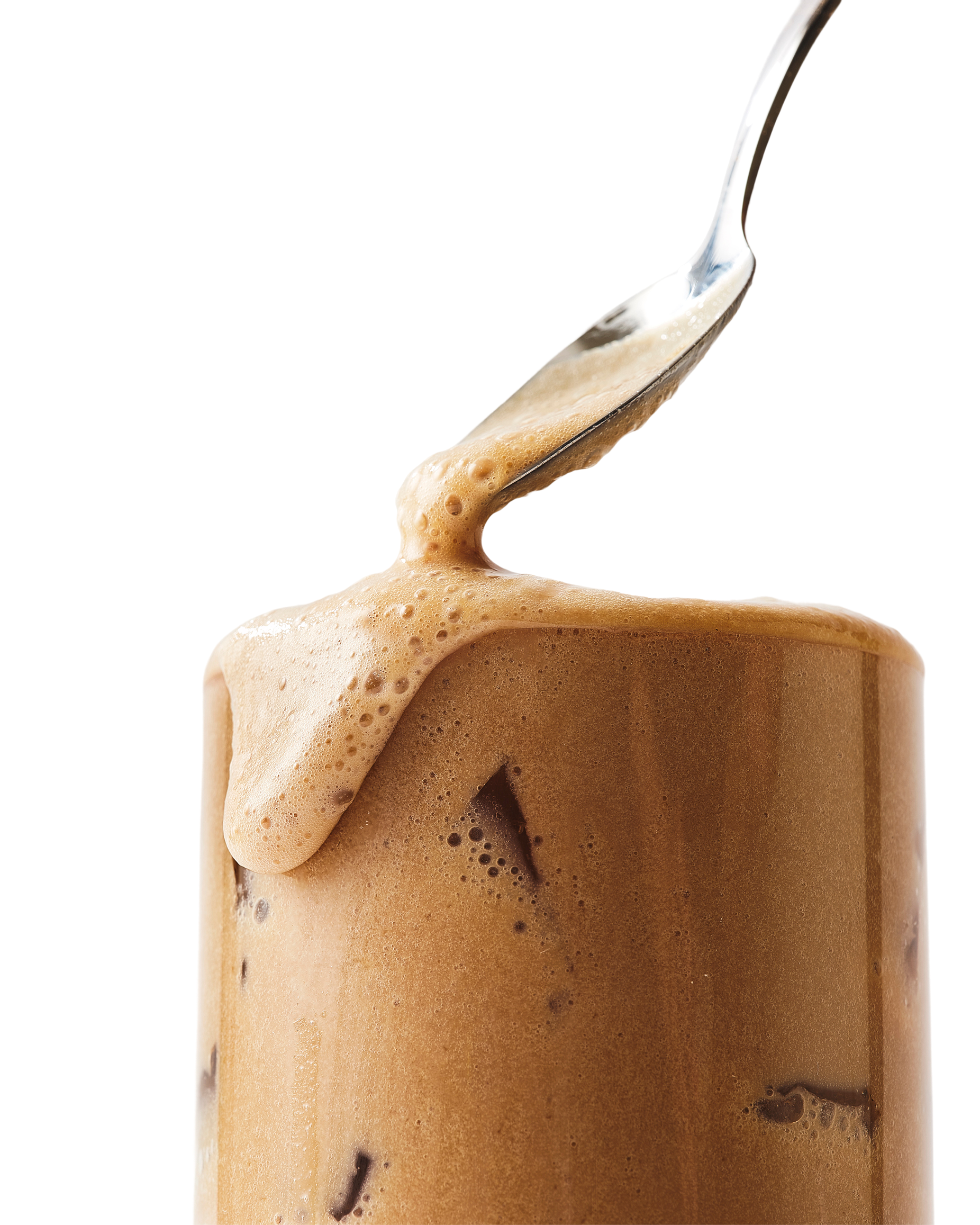A spoon drizzles frothy foam over an iced coffee in a clear glass.