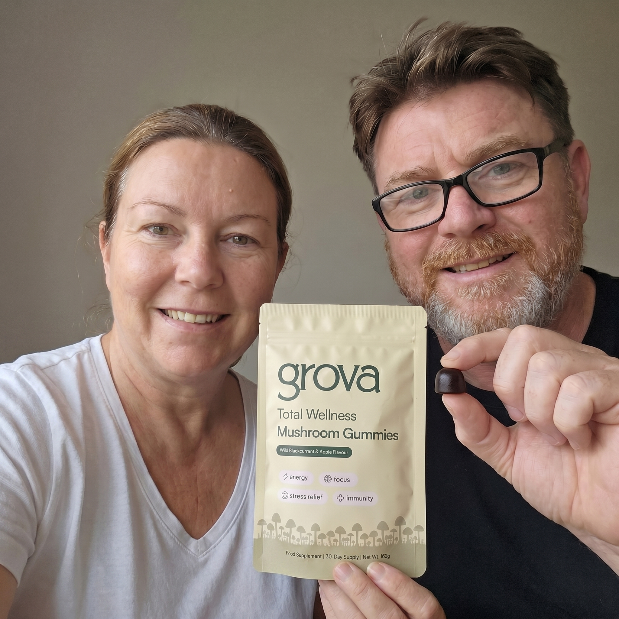 A smiling man and woman hold up a pouch of Grova Mushroom Gummies and one individual gummy.