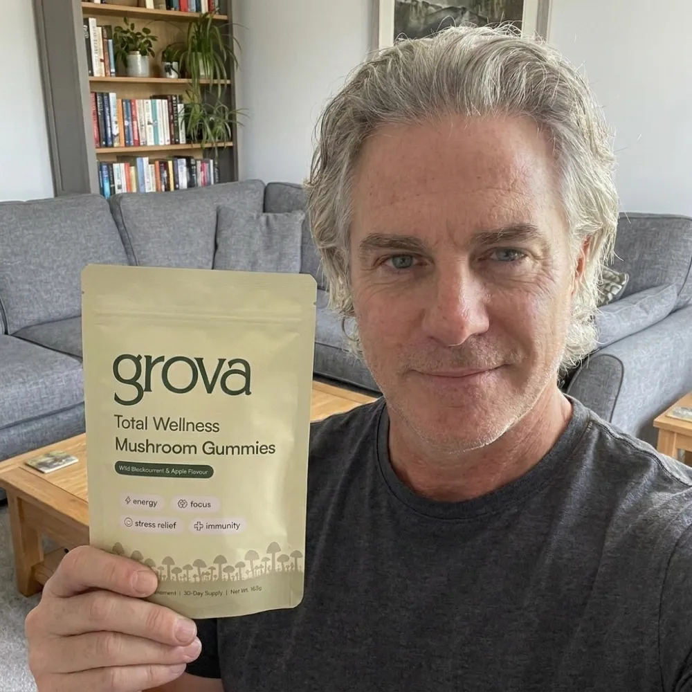A man with gray hair holds a pouch of grova Total Wellness Mushroom Gummies in a living room.