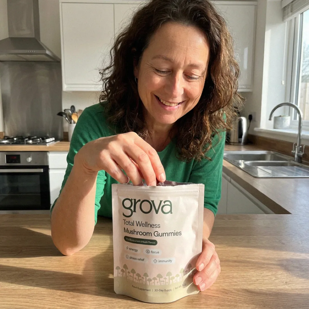 Smiling woman holding a pack of Grova Total Wellness Mushroom Gummies in a kitchen.