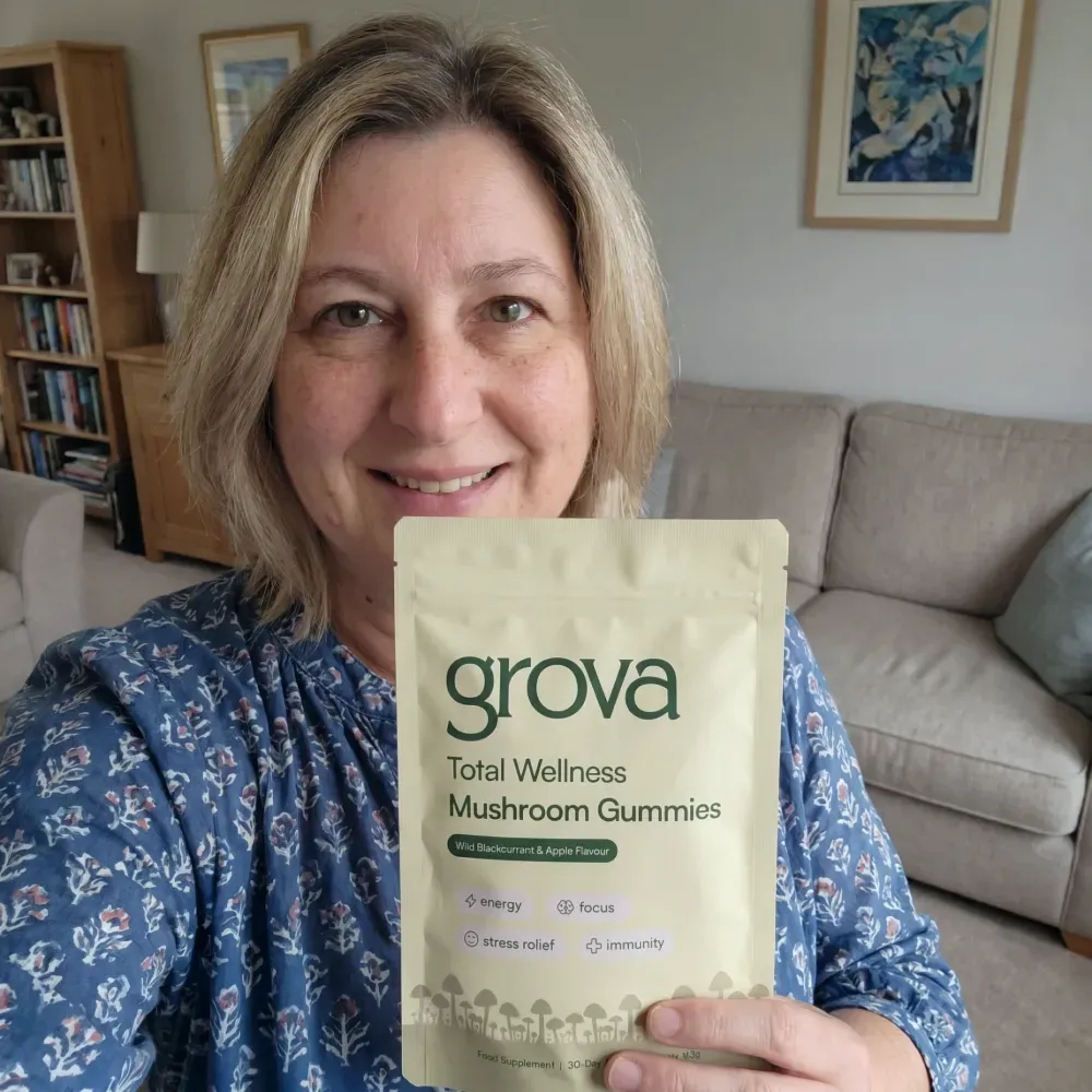 A smiling woman holds a pouch of grova Total Wellness Mushroom Gummies in a living room.