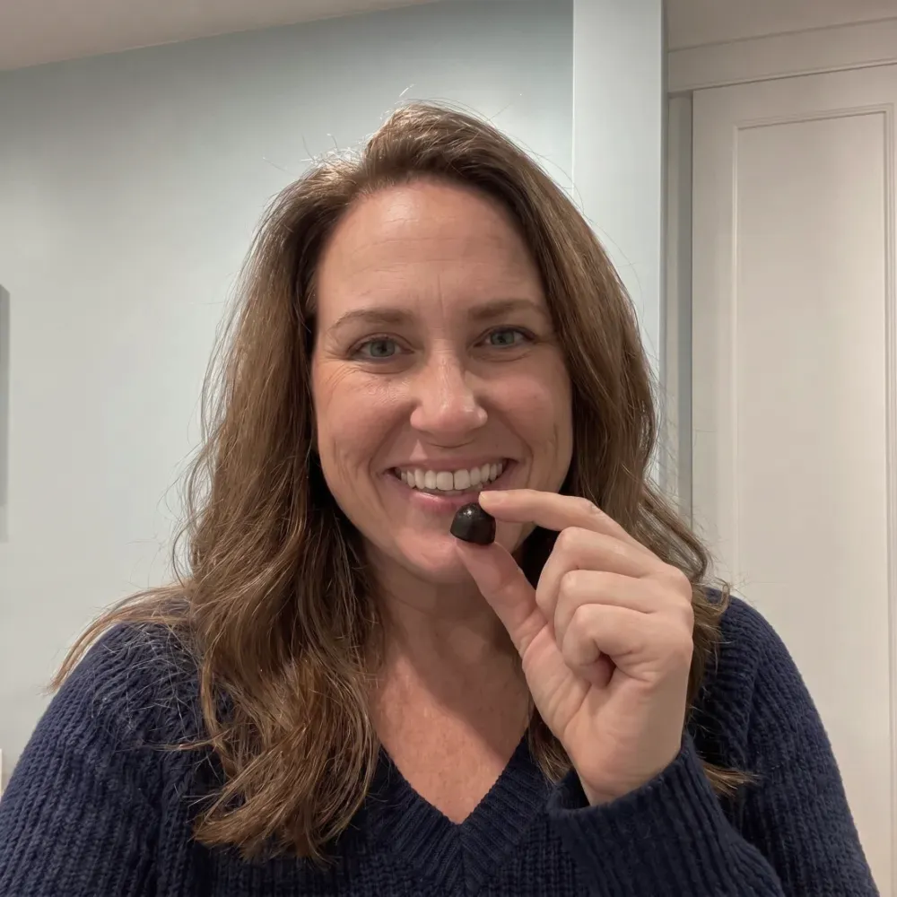 A smiling woman in a blue sweater holding up a small, dark, round piece of candy.