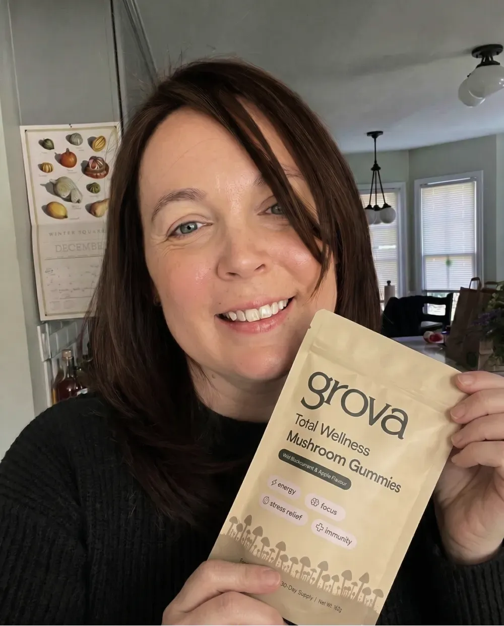 A smiling woman holds up a pouch of grova Total Wellness Mushroom Gummies.
