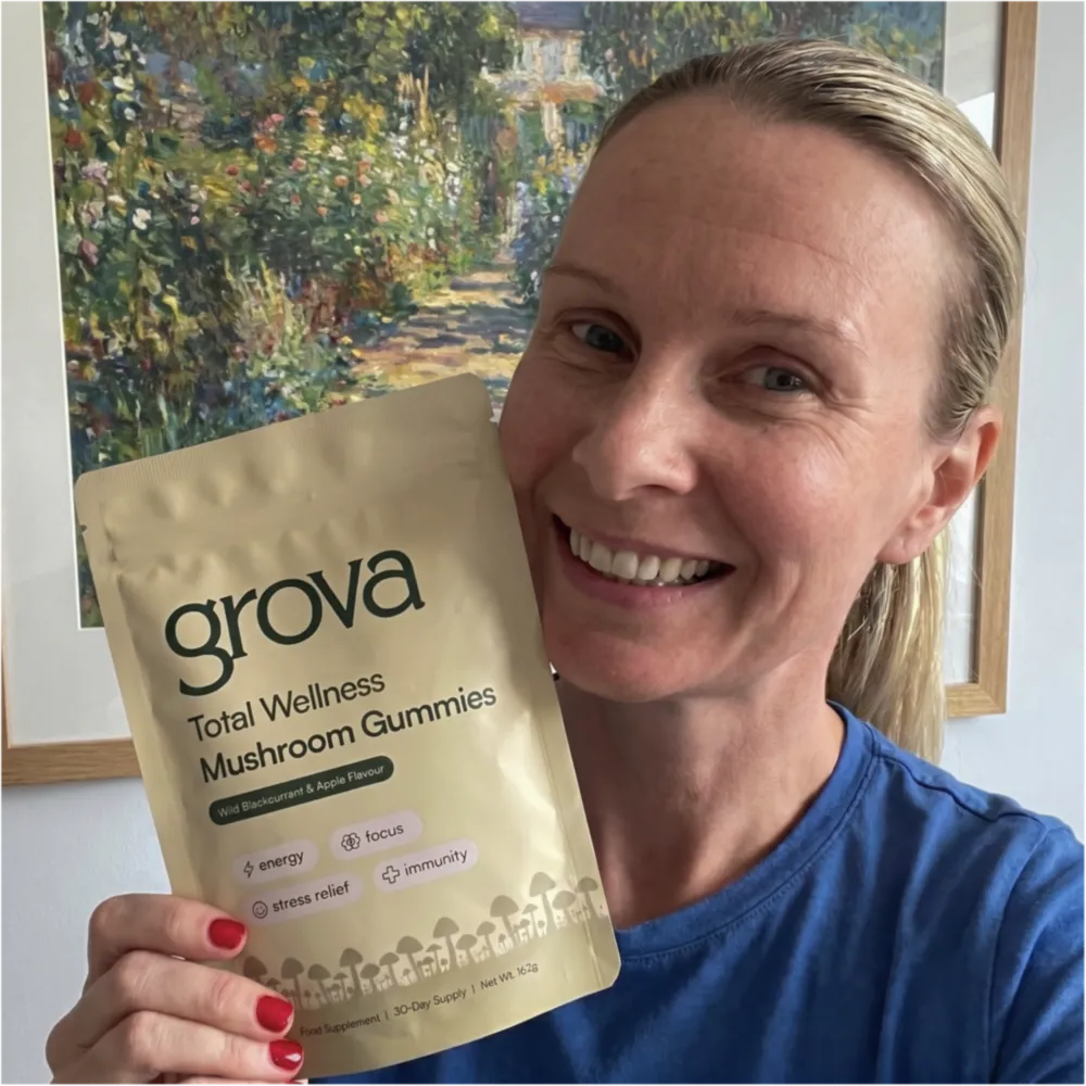 A smiling woman in a blue shirt holds up a pouch of Grova Total Wellness Mushroom Gummies.