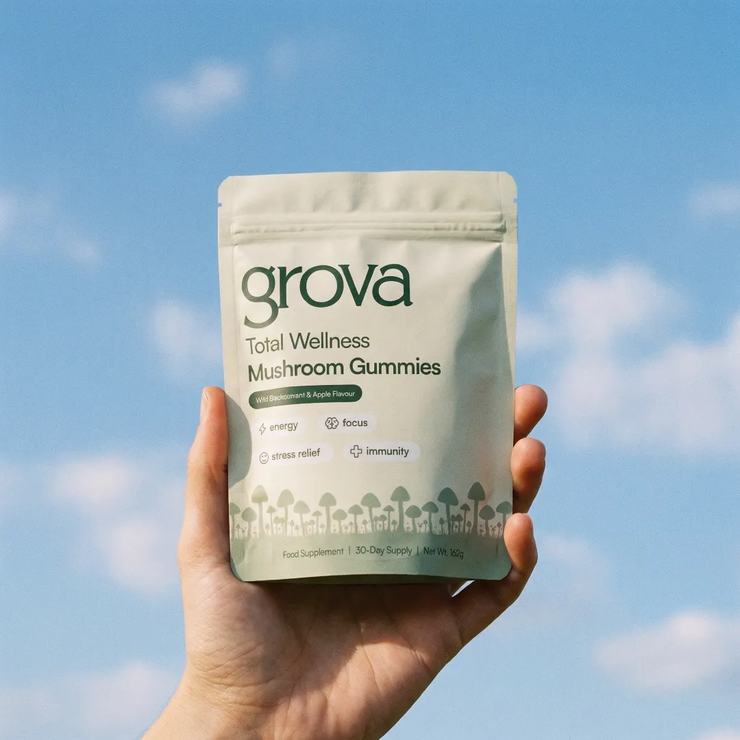 A hand holds a pouch of Grova Total Wellness Mushroom Gummies against a blue sky.