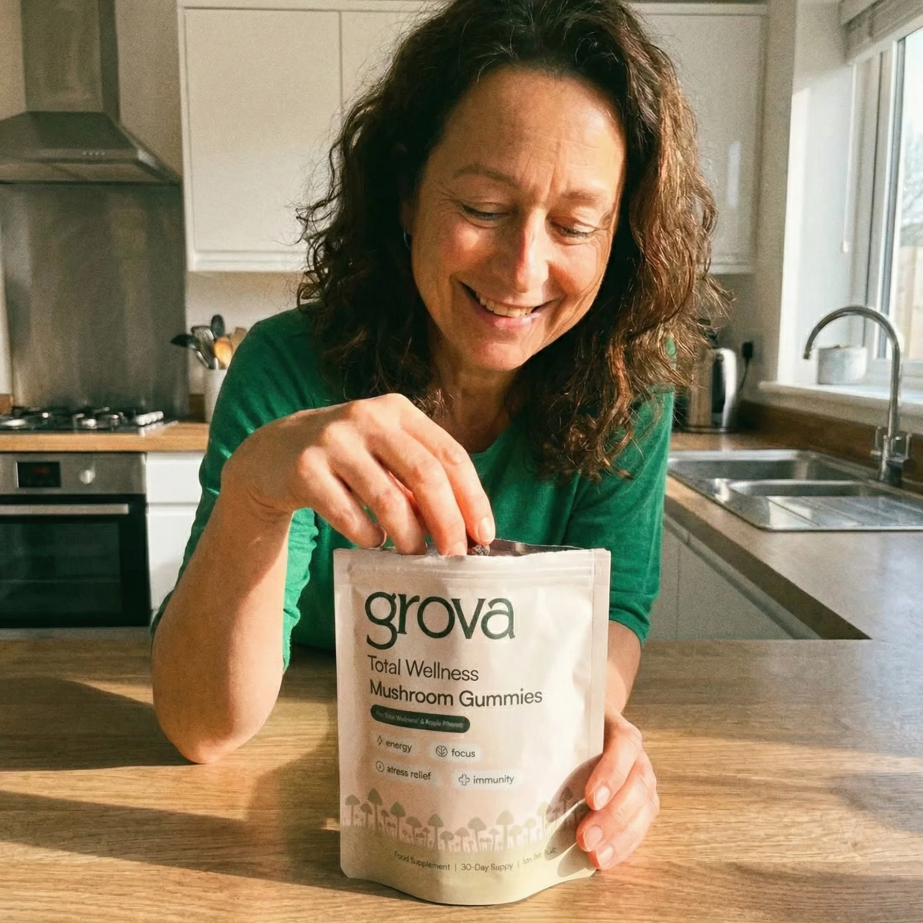 A smiling woman in a green shirt reaches into a pouch of grova Mushroom Gummies in a kitchen.