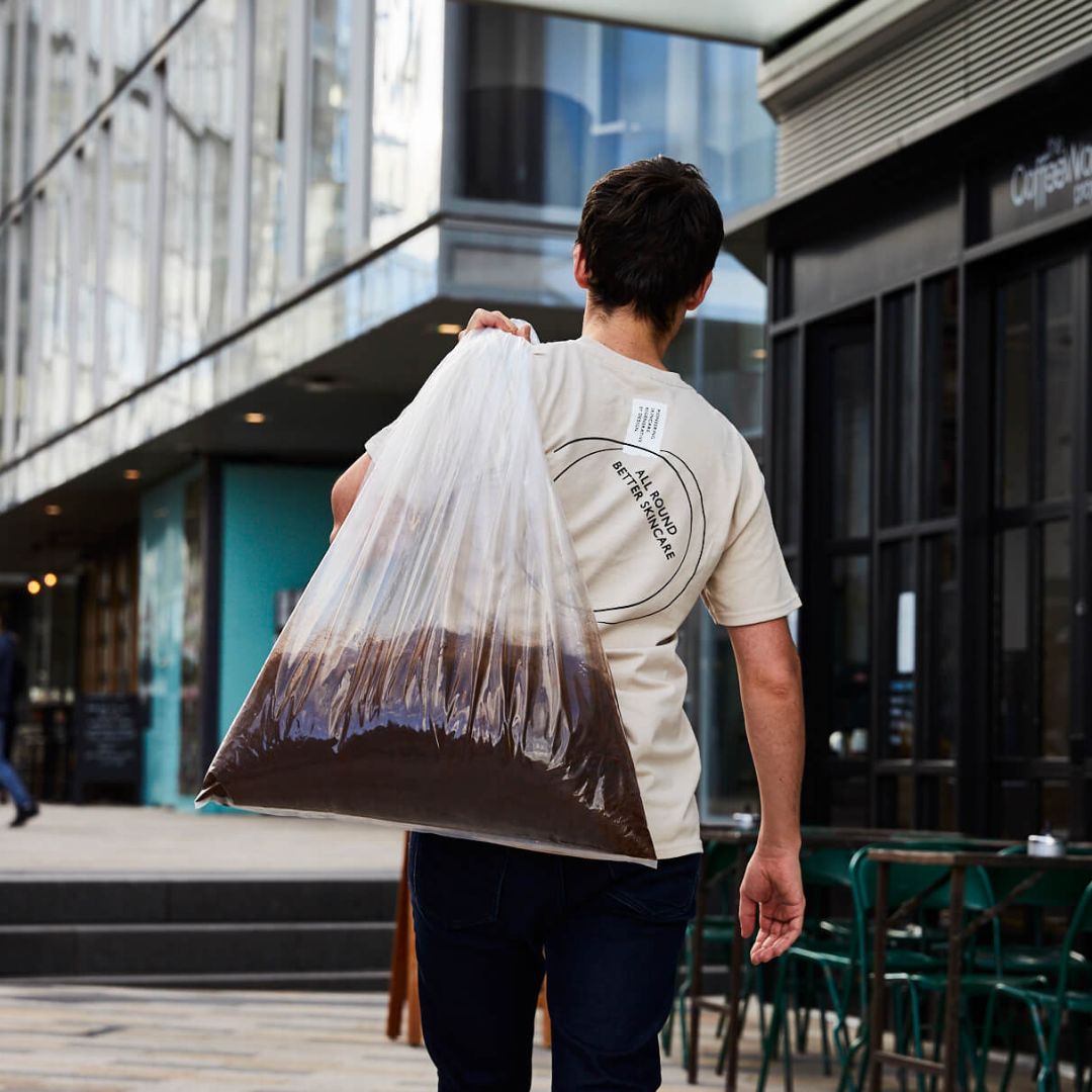 A person seen from behind carries a large, clear bag of used coffee grounds over their shoulder.