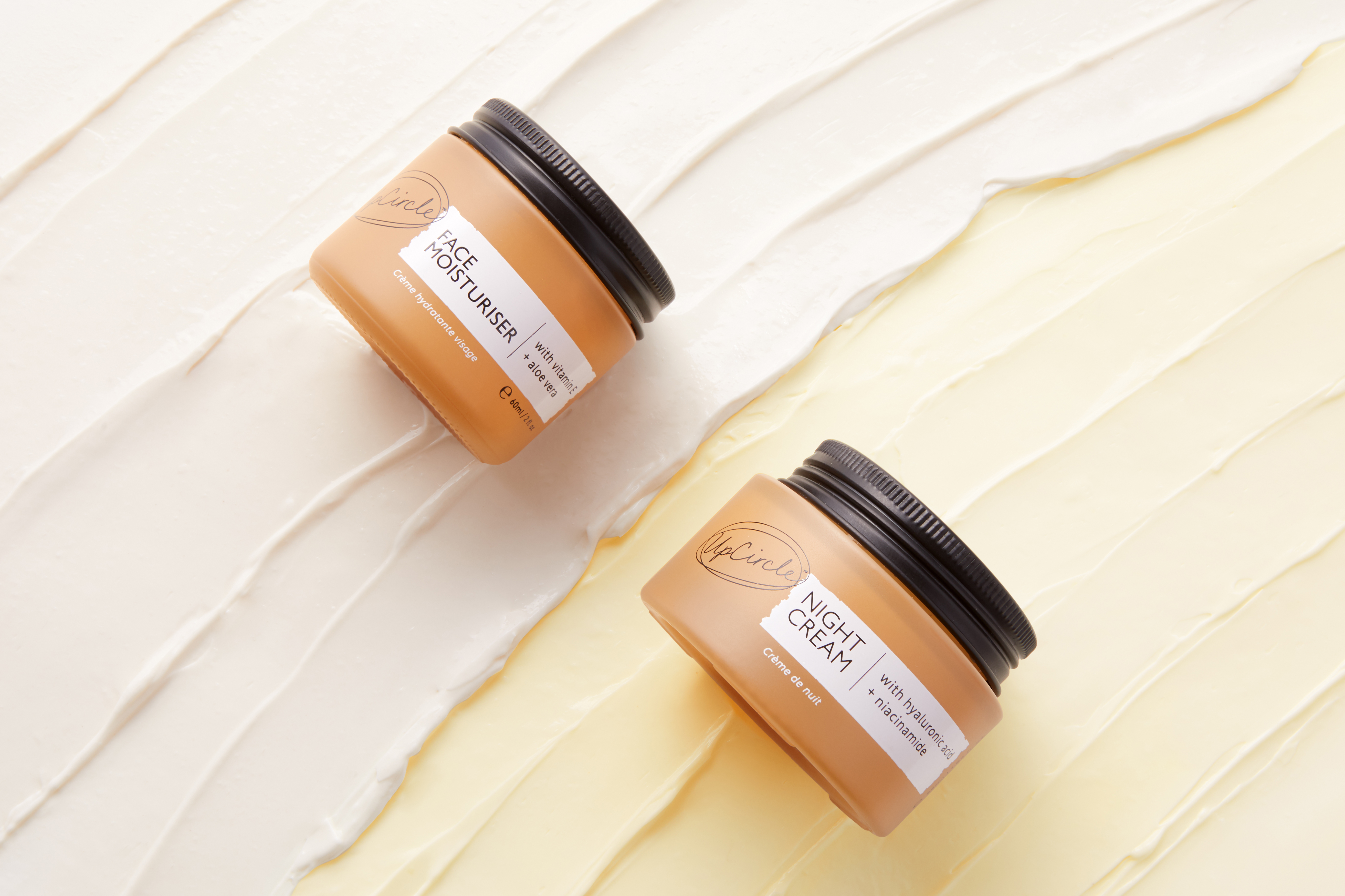 Two jars of UpCircle moisturiser and night cream lying diagonally on a background of smeared white and yellow cream.