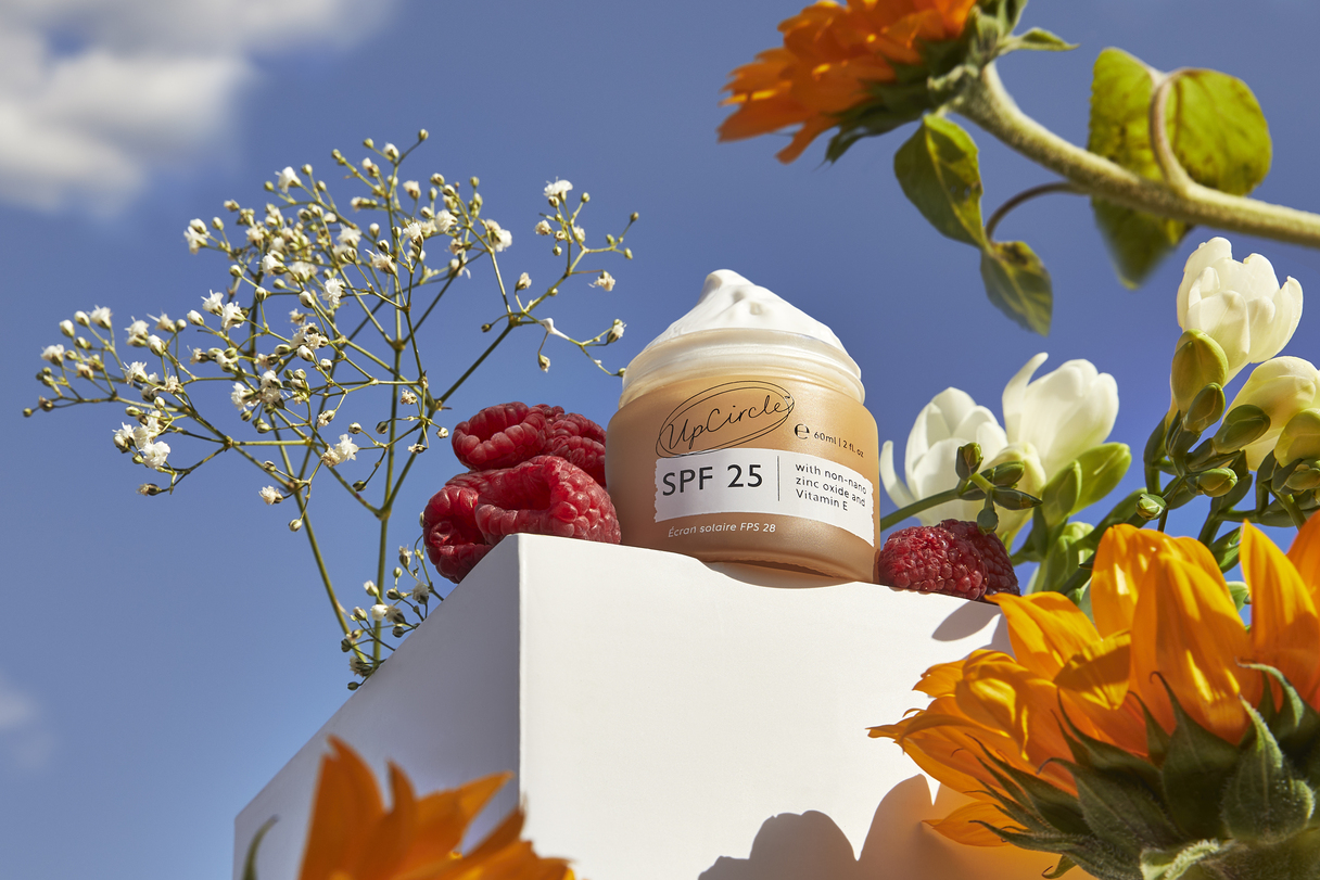 A jar of UpCircle SPF 25 cream sits on a white block, surrounded by flowers and raspberries.