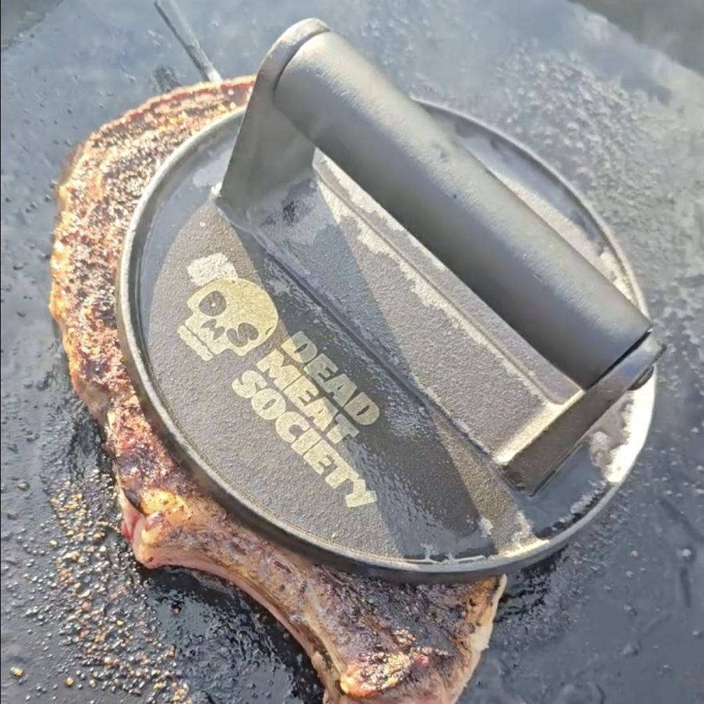A steak is seared on a griddle using a round cast-iron press with a 'Dead Meat Society' logo.