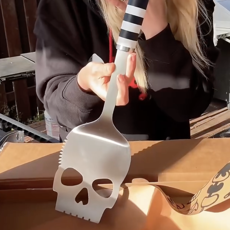 A person holds up a large, metal barbecue spatula with the head shaped like a skull.