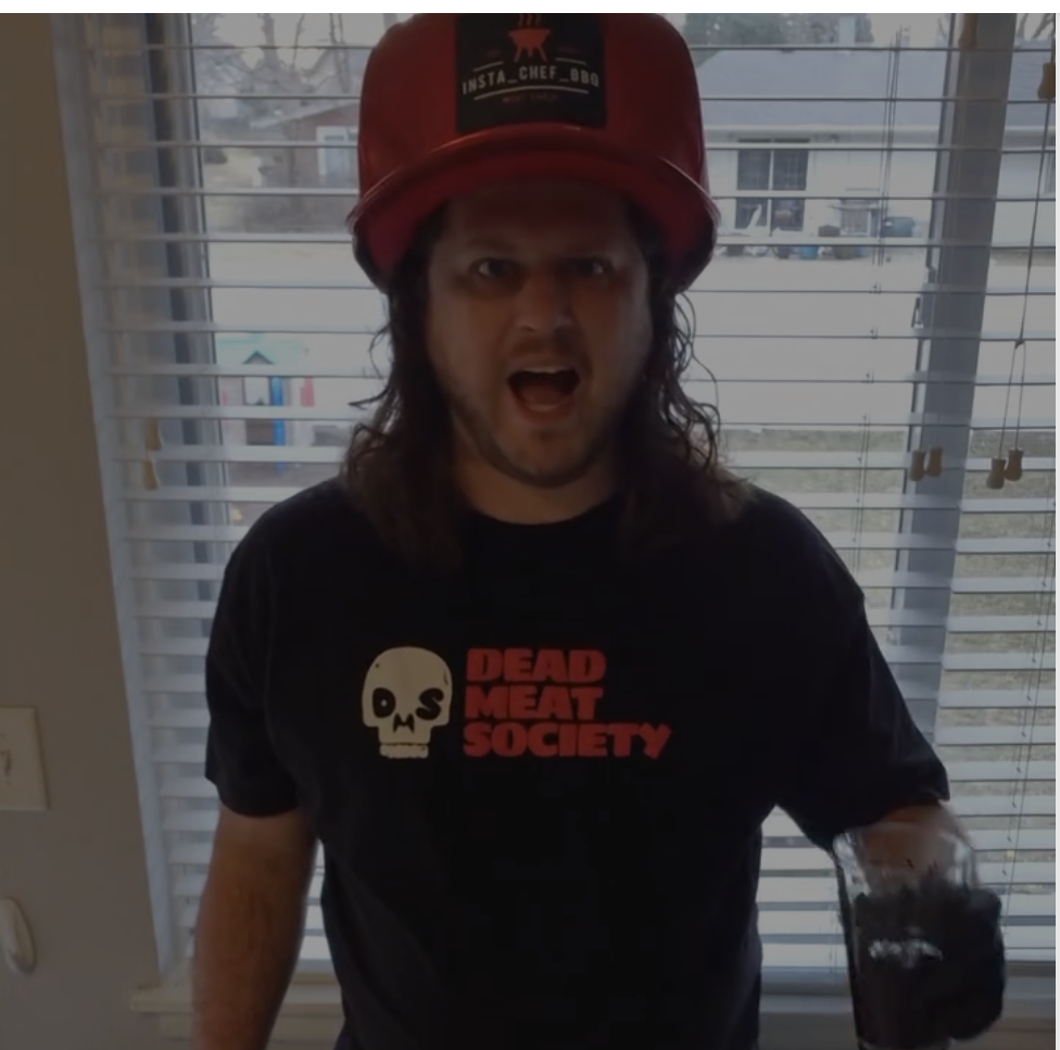 A man with long hair in a red hat and a black graphic t-shirt yells at the camera.