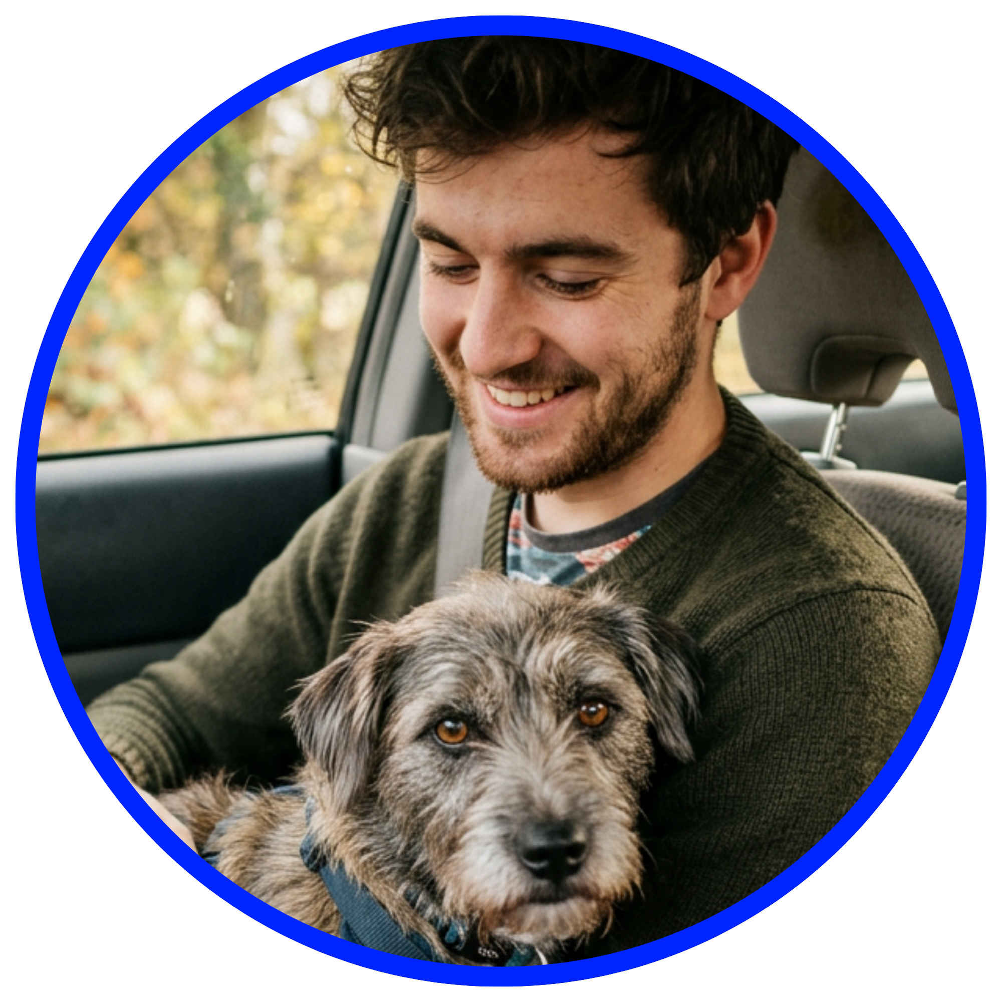 A smiling young man with a beard holds a scruffy terrier-type dog in his lap inside a car.
