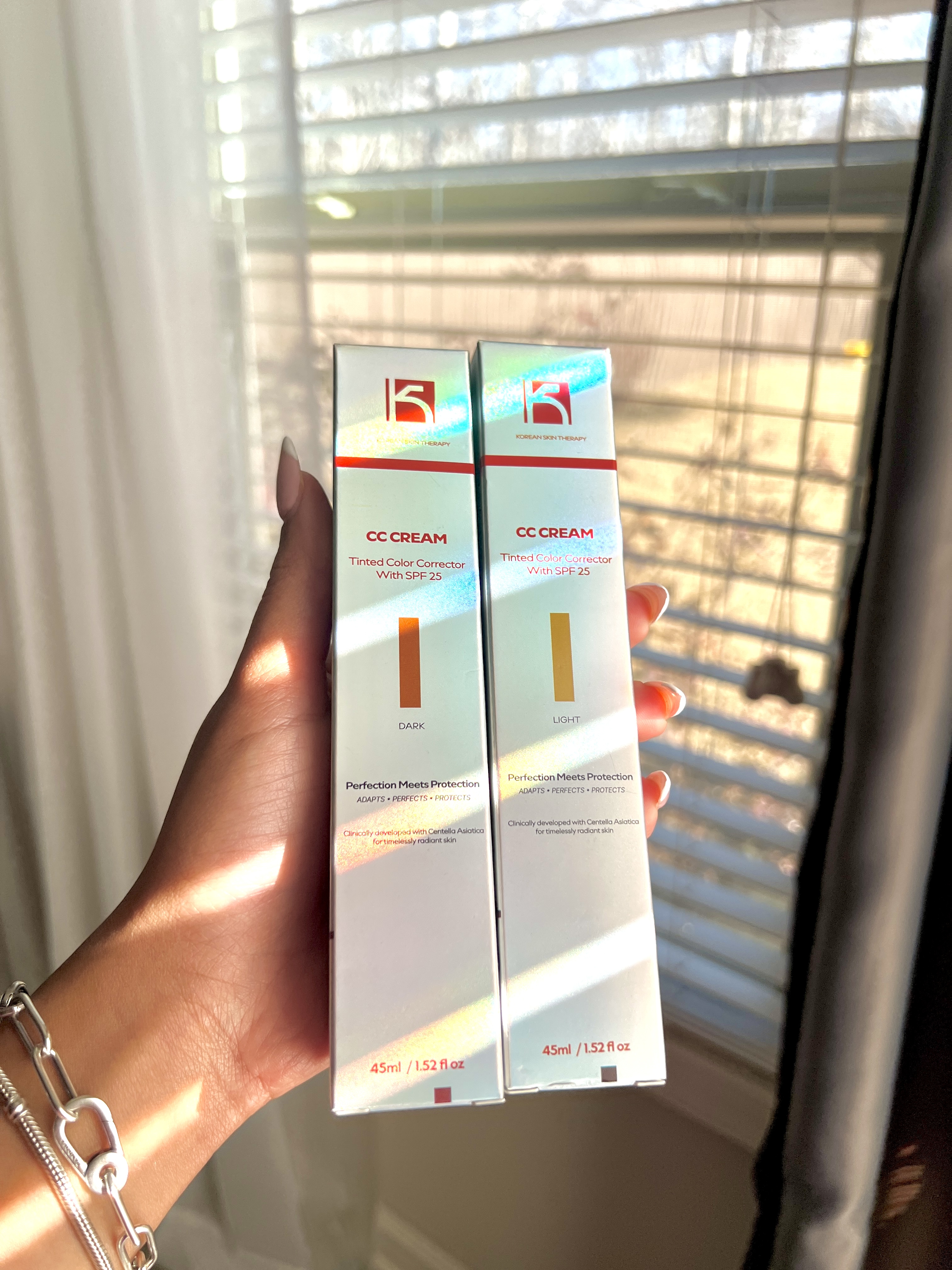 Two boxes of CC cream held in front of a window with blinds.