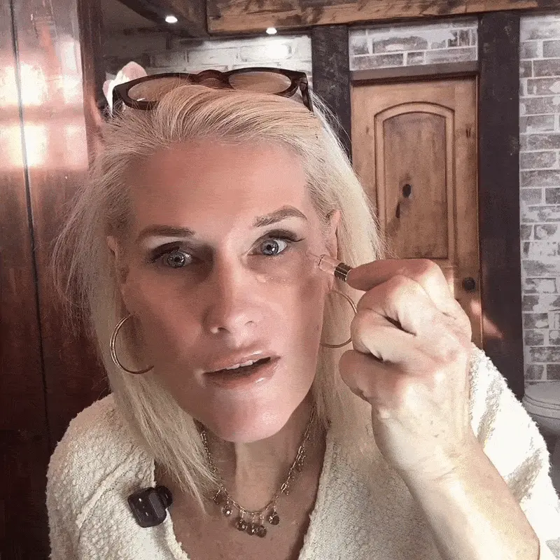 A woman with blonde hair applies a serum or cosmetic product under her right eye with an applicator.