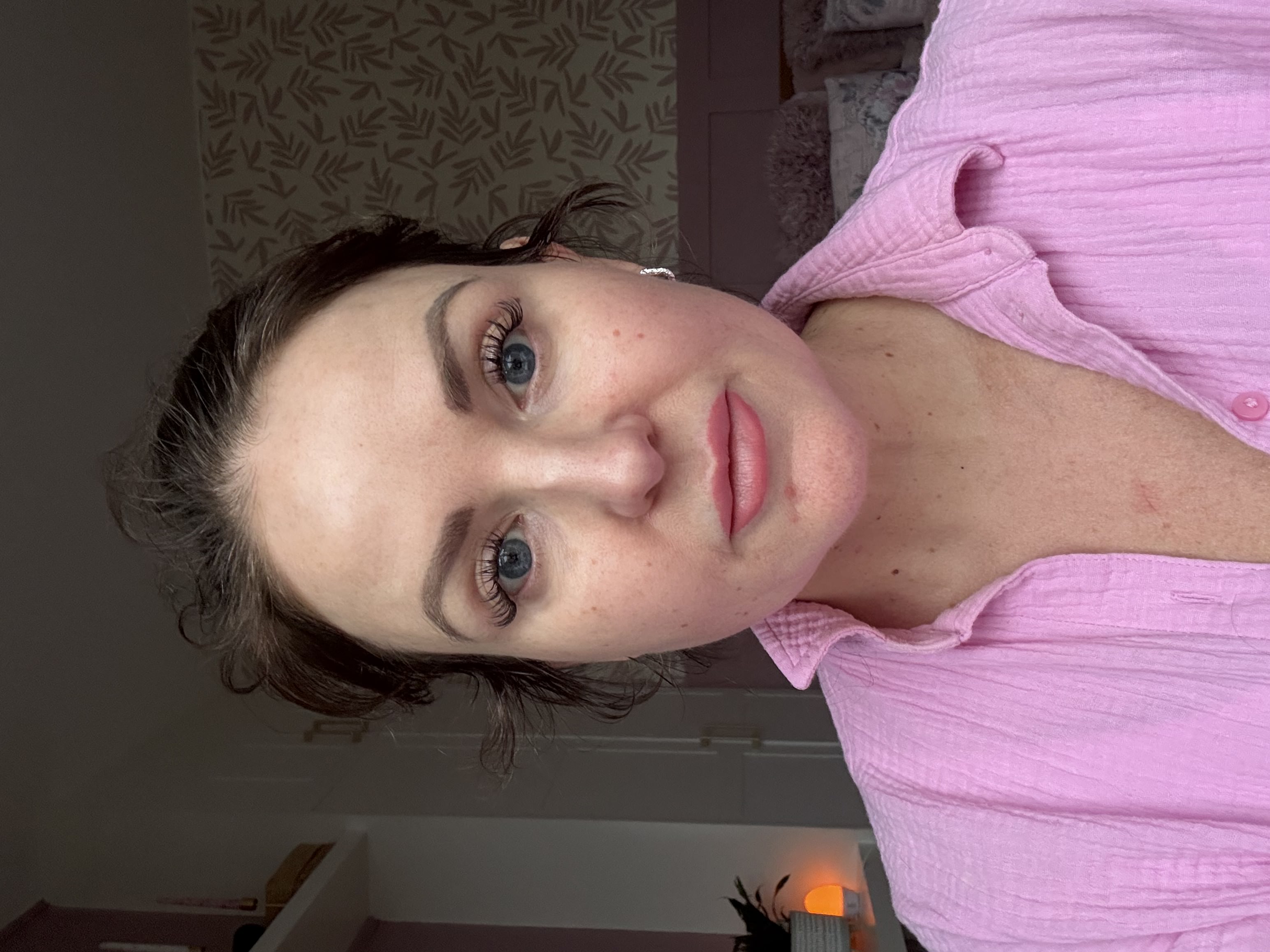 Person wearing a pink shirt, indoors, with a patterned wall in the background.
