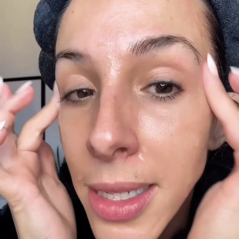 A woman with a black headband on pats a clear liquid onto her face with her fingers.