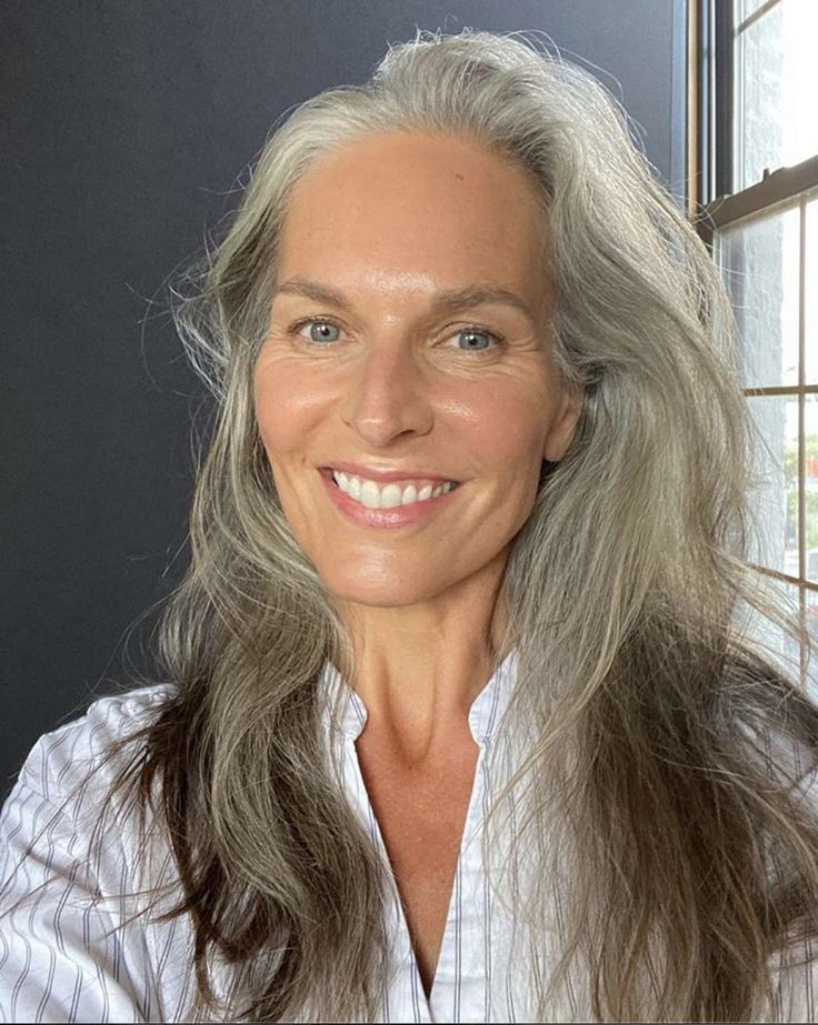 Smiling person with long gray hair and a white shirt near a window.