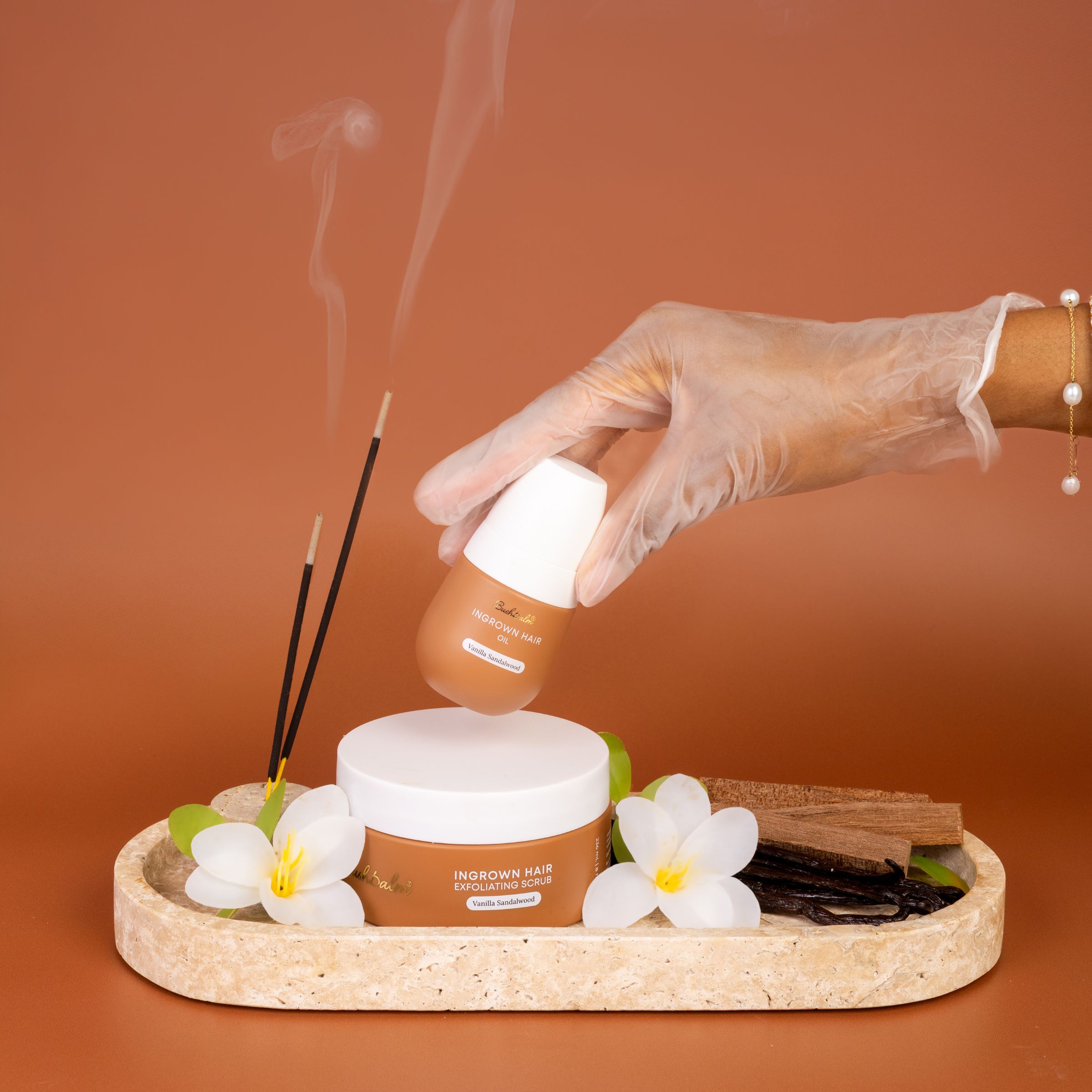Hand holding ingrown hair oil above scrub, with flowers and incense on a tray.