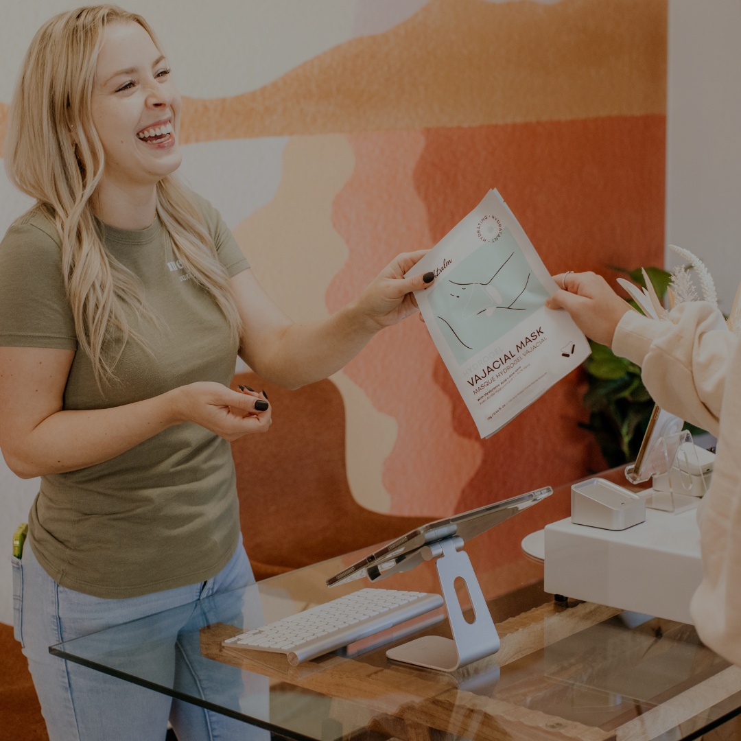 A smiling woman behind a counter hands a skincare product package to another person.