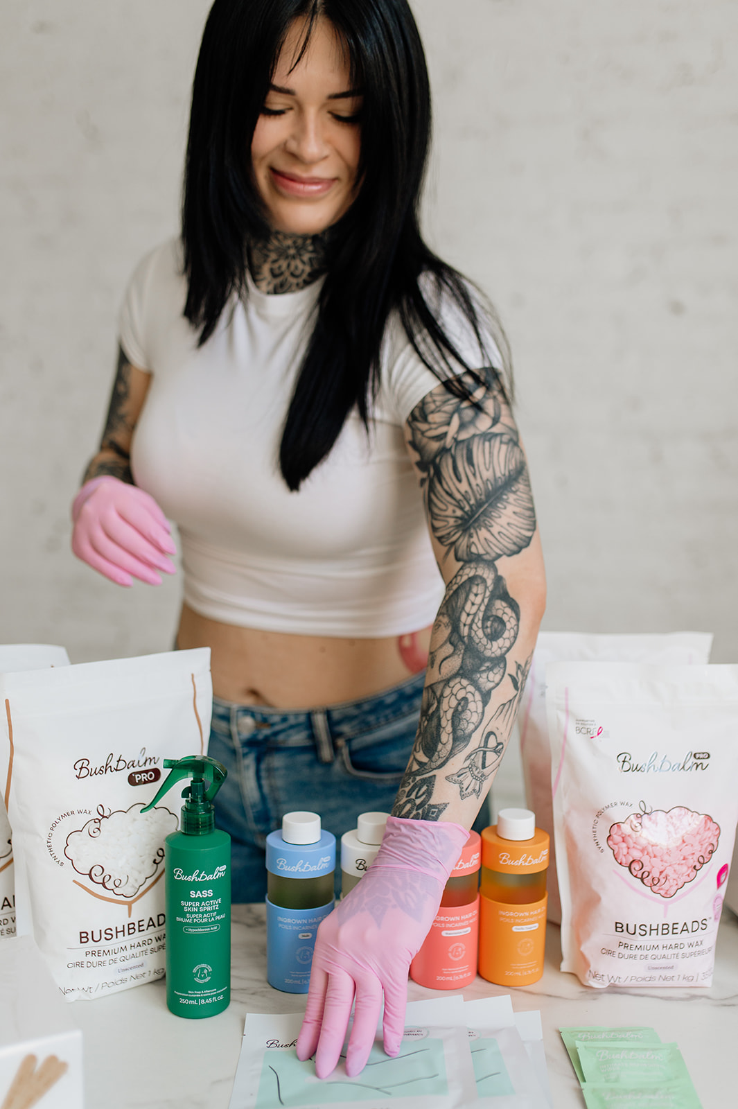 A tattooed woman wearing pink gloves arranges Bushbalm skincare and waxing products on a table.