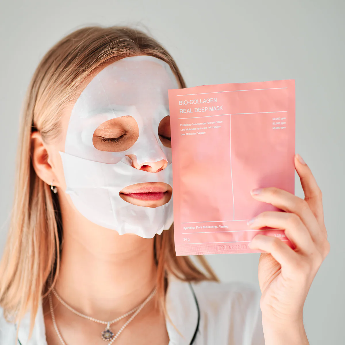 Person wearing a sheet mask holding a pink skincare product package.