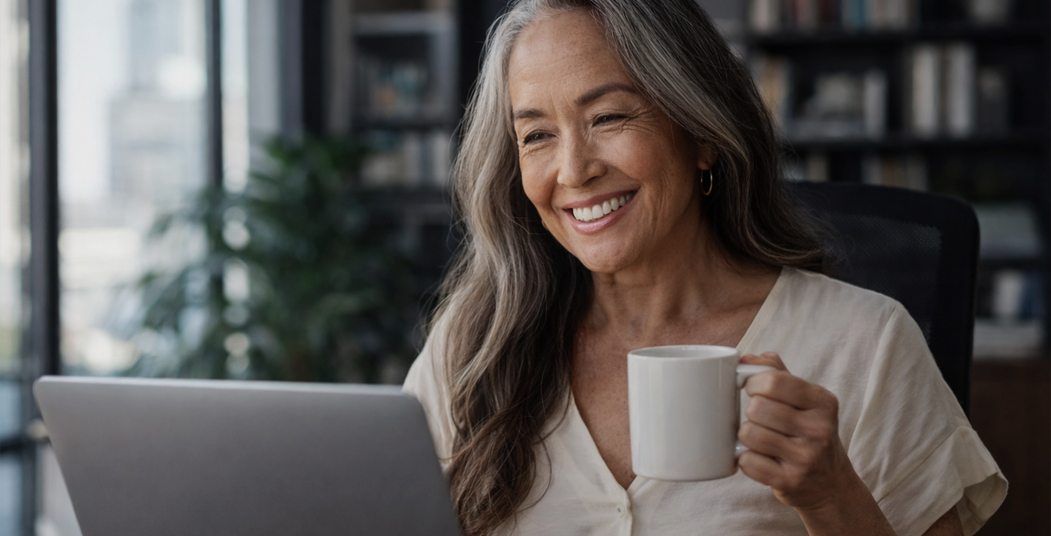 A smiling older woman with long gray hair holds a mug while looking at a laptop.