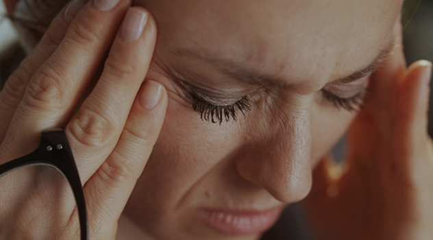 A close-up of a woman with her eyes closed, pressing her fingers to her temples while holding glasses.