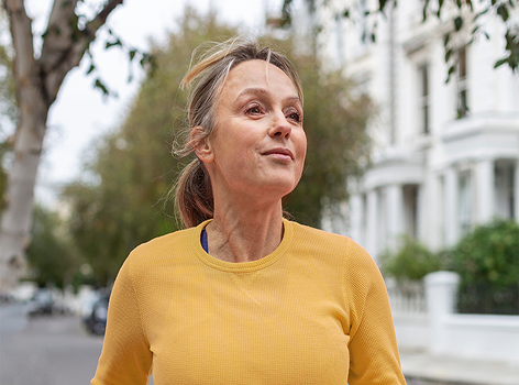 A middle-aged woman in a yellow shirt stands outdoors on a tree-lined street.