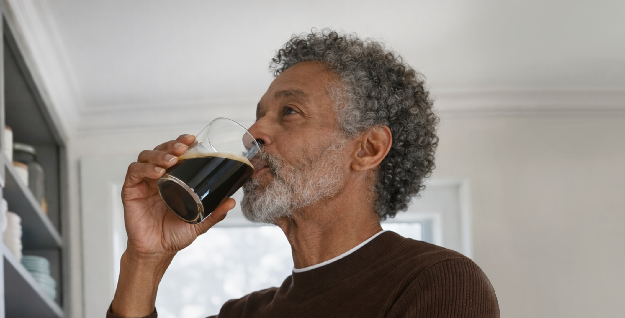 An older man with gray curly hair and a gray beard drinks a dark beverage from a glass.