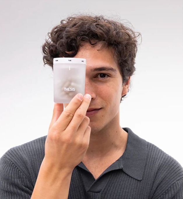A man in a grey polo shirt holds a Thesis pill organizer over his eye.