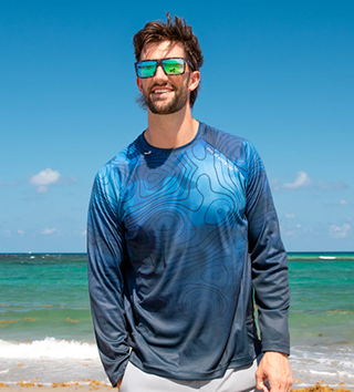Man in sunglasses and blue shirt standing on a beach with ocean in the background.