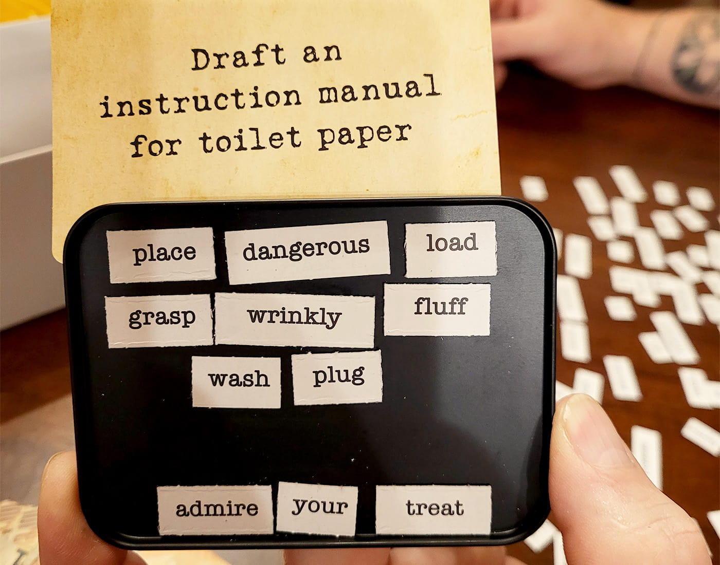 A game prompt card reads 'Draft an instruction manual for toilet paper' above a tray of words.